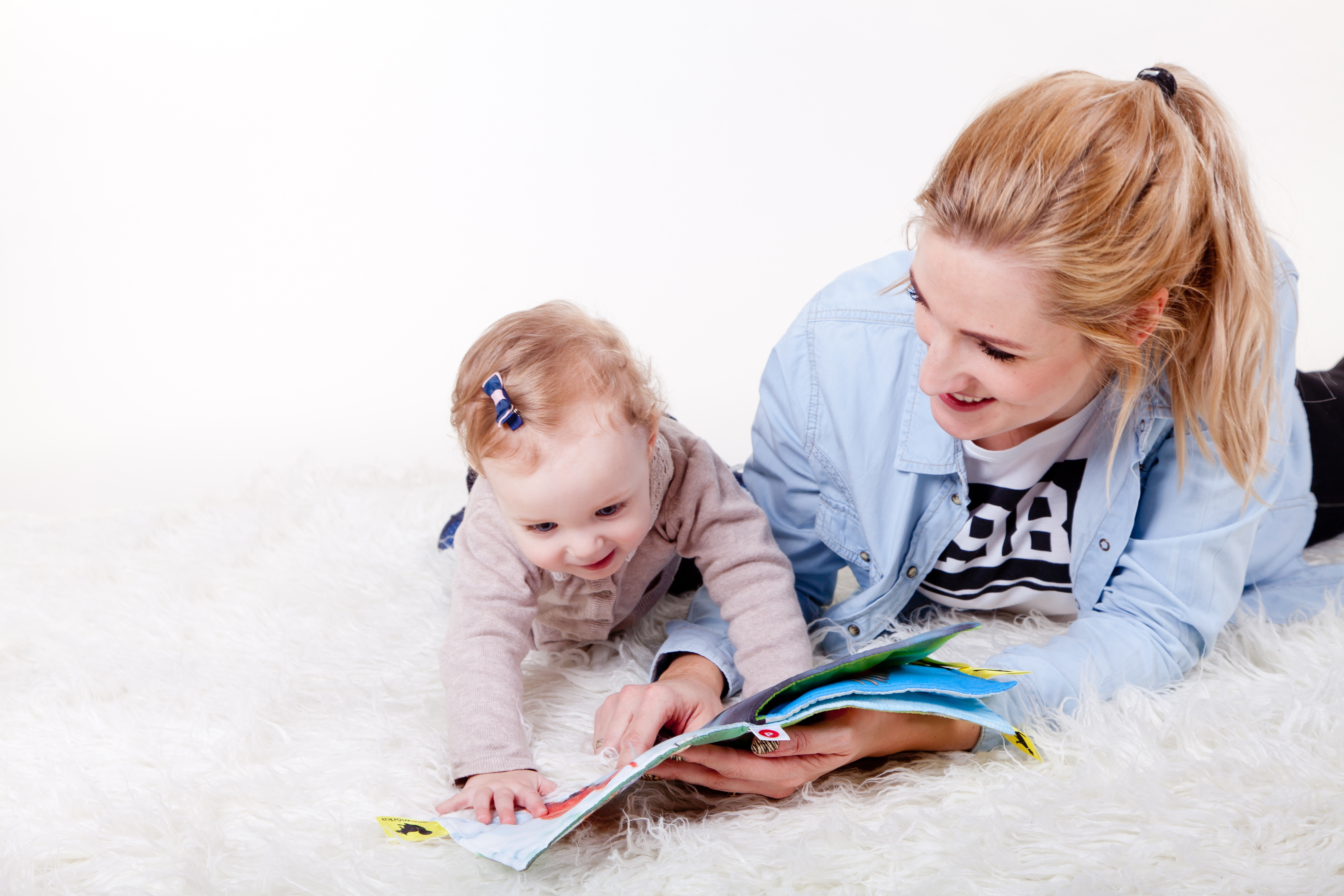 Baby and mother lying on a white rug reading a book together.